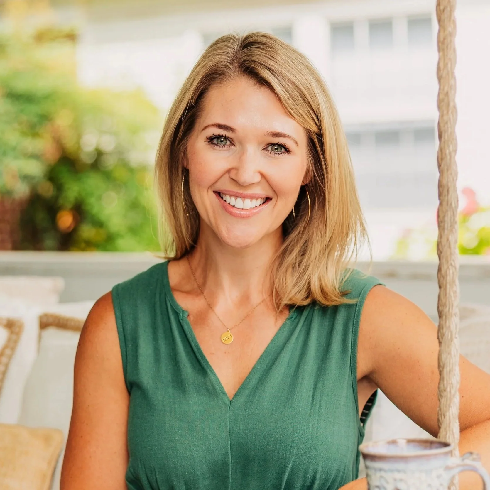 Close-up of a smiling woman with blonde hair, wearing a green sleeveless top, and sitting on a wooden swing with rope, outdoors with blurred greenery and house in the background.