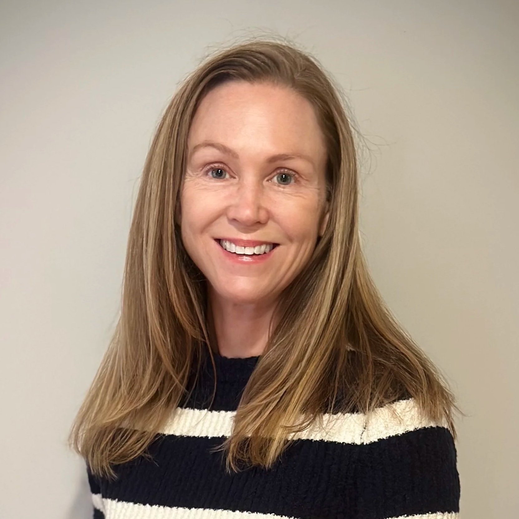 A smiling woman with long, light brown hair, wearing a black and white striped sweater, standing against a plain, light-colored wall.