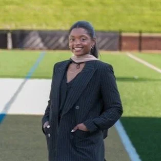 Young woman with braided hair in a black pinstripe suit smiling outdoors on a sports field.
