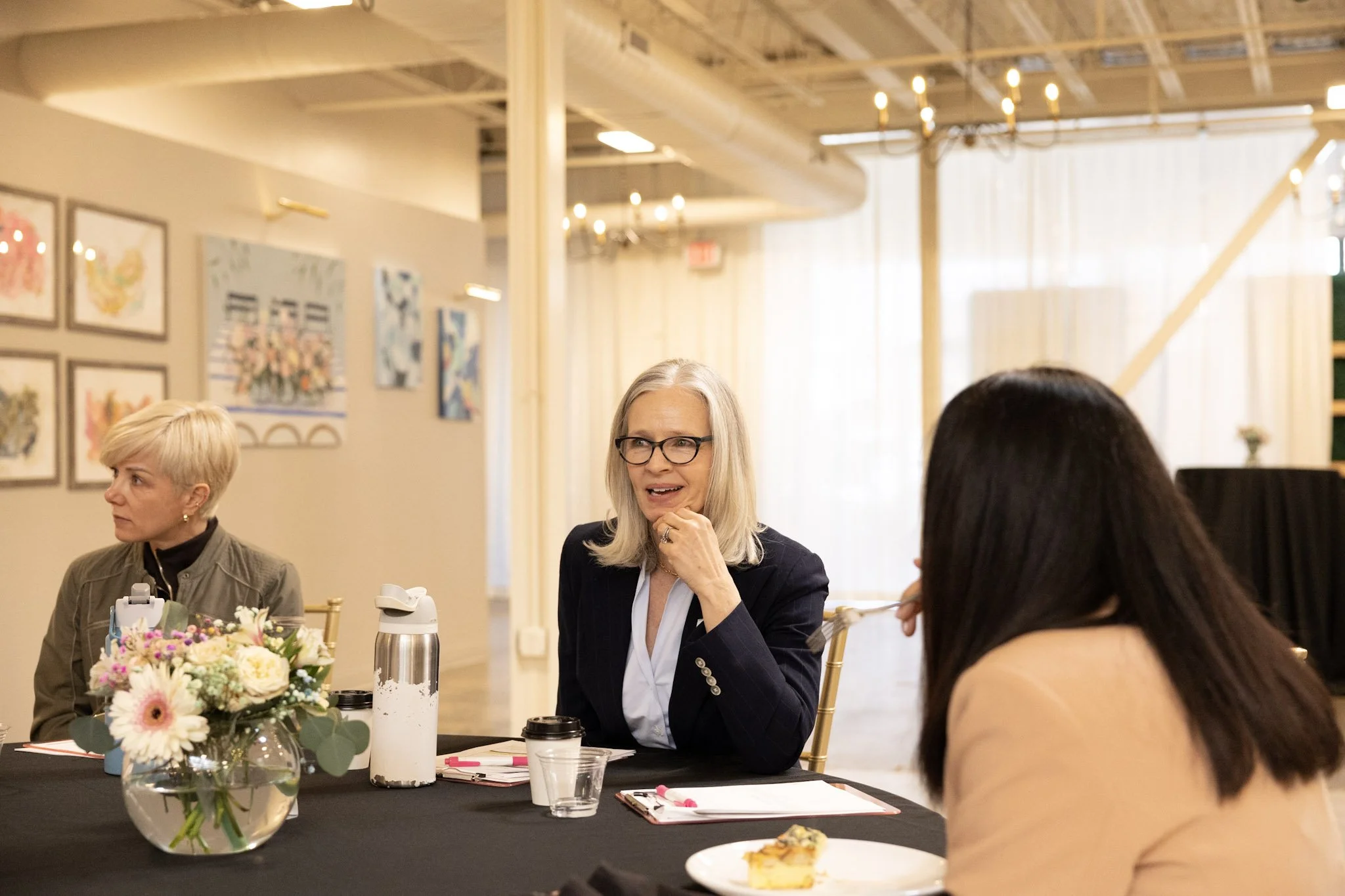 A group of women seated at a table during a meeting or discussion in a well-lit, decorated room with wall art and hanging lights.