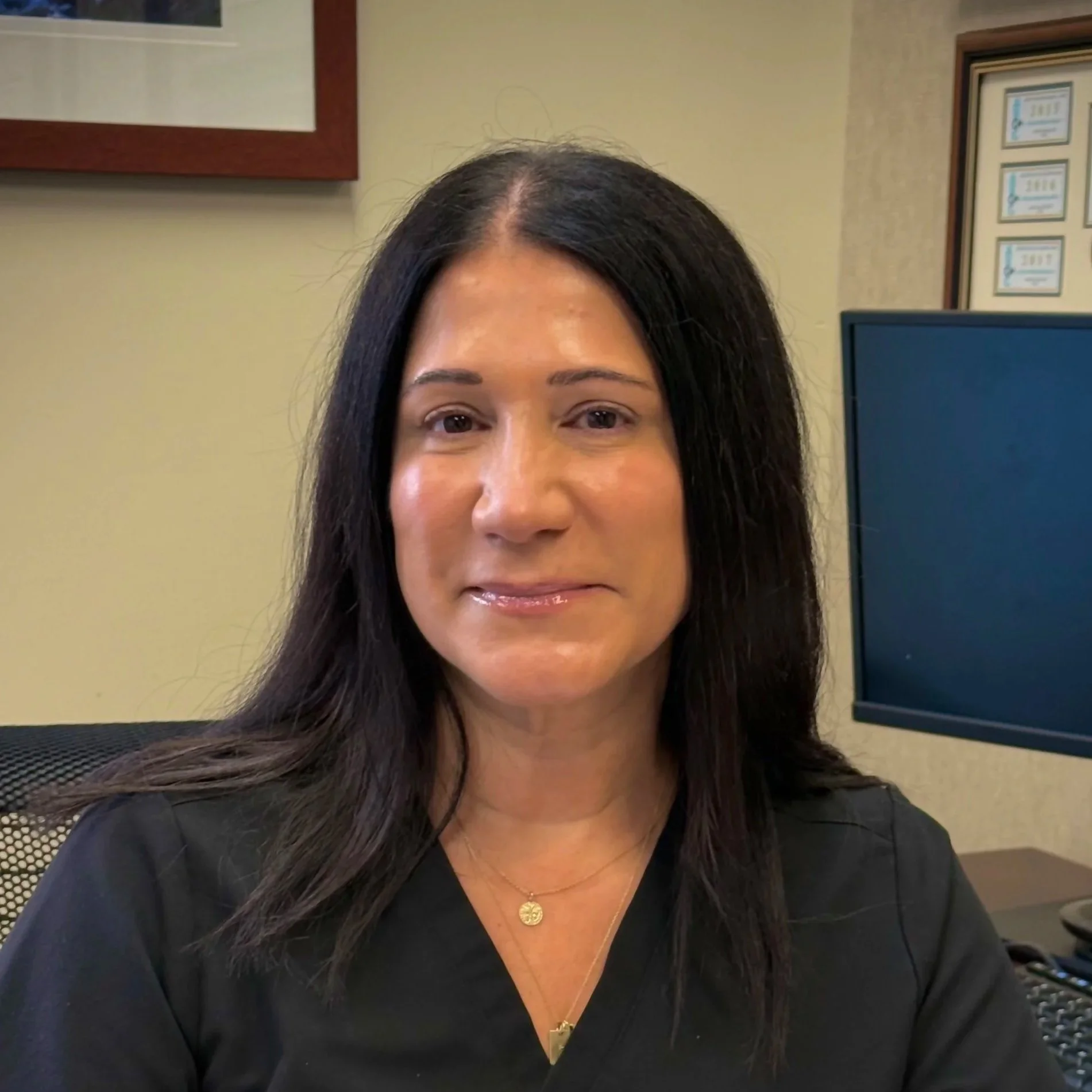 Close-up of a woman with long black hair, wearing a black top, gold jewelry, and a smile in an office setting with framed certificates on the wall.