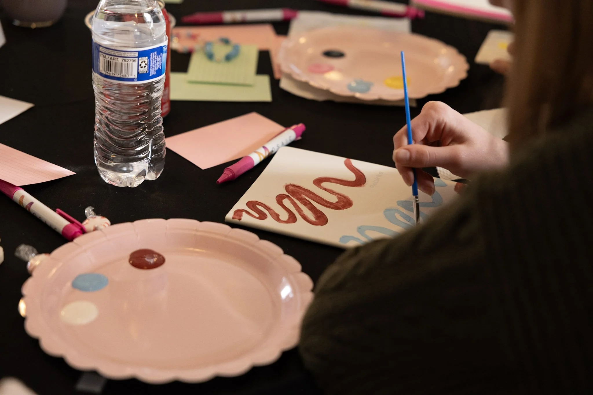 Person painting on a white paper with red and blue paint, surrounded by pink paper plates, markers, a water bottle, and decorative heart-shaped pink plates with paint on them, on a black table.