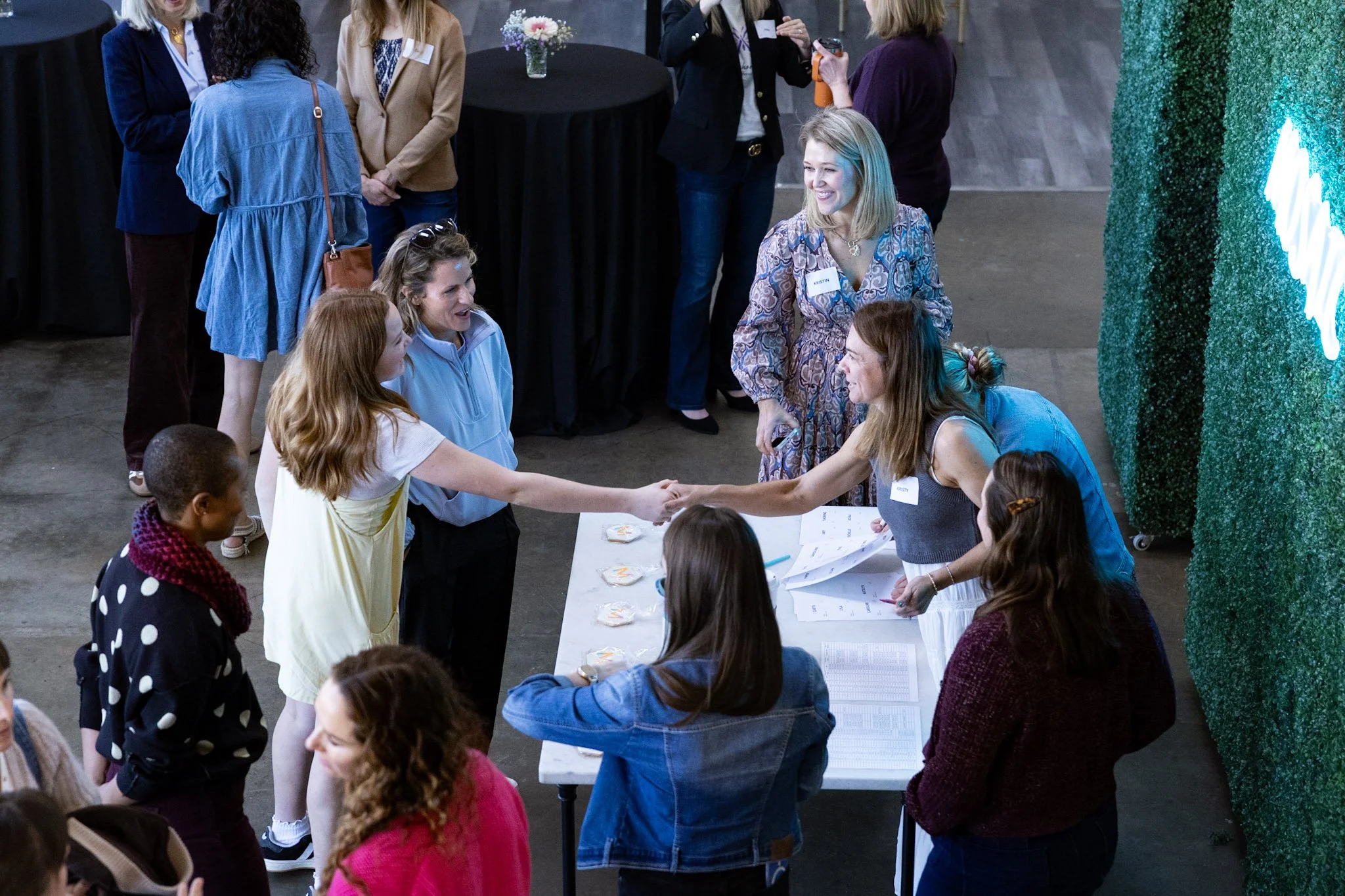 People gathered around a table at a networking event or conference, shaking hands and engaging in conversation. The scene includes women standing and talking, and there are name tags visible on some individuals.
