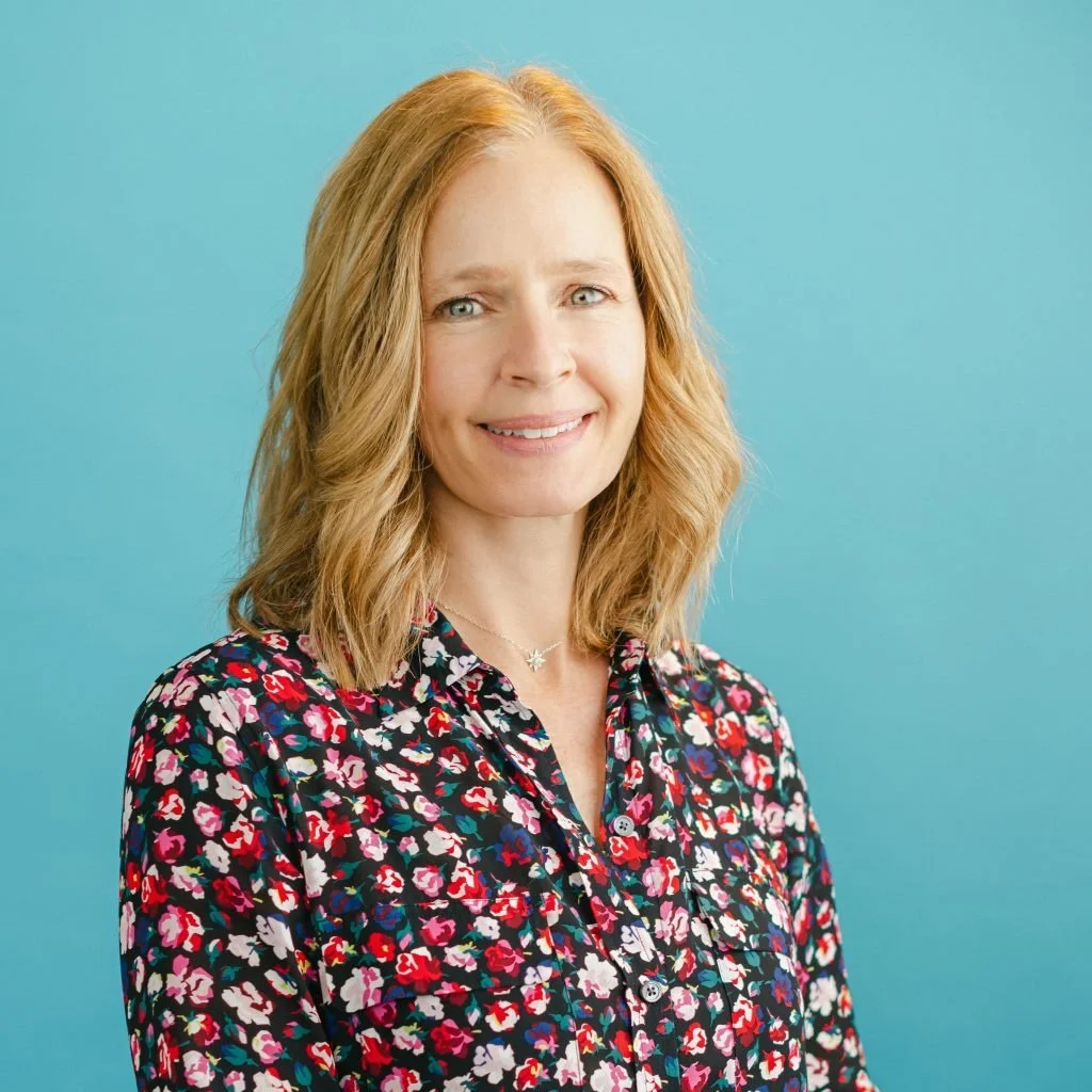 A woman with shoulder-length blonde hair, smiling, wearing a colorful floral blouse, standing against a light blue background.