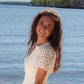 A smiling woman with curly hair wearing a white dress, standing near a body of water with trees in the background.