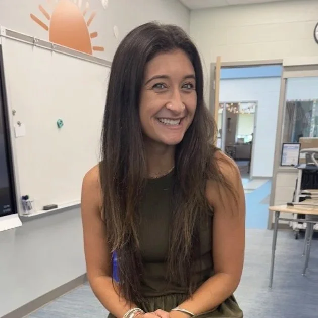 A young woman with long brown hair sitting and smiling in a classroom or office setting.