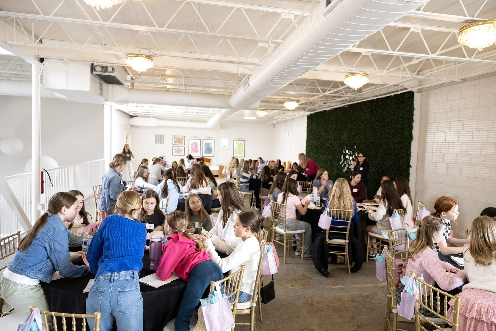 Group of young girls sitting at tables during a daytime event in a spacious modern indoor venue with white walls and green wall decor.