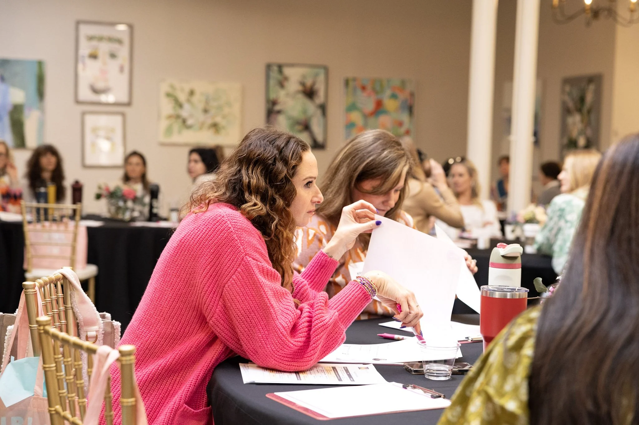 Women sitting at a table in an art gallery or meeting room, engaged in discussion, with artwork on the walls in the background.