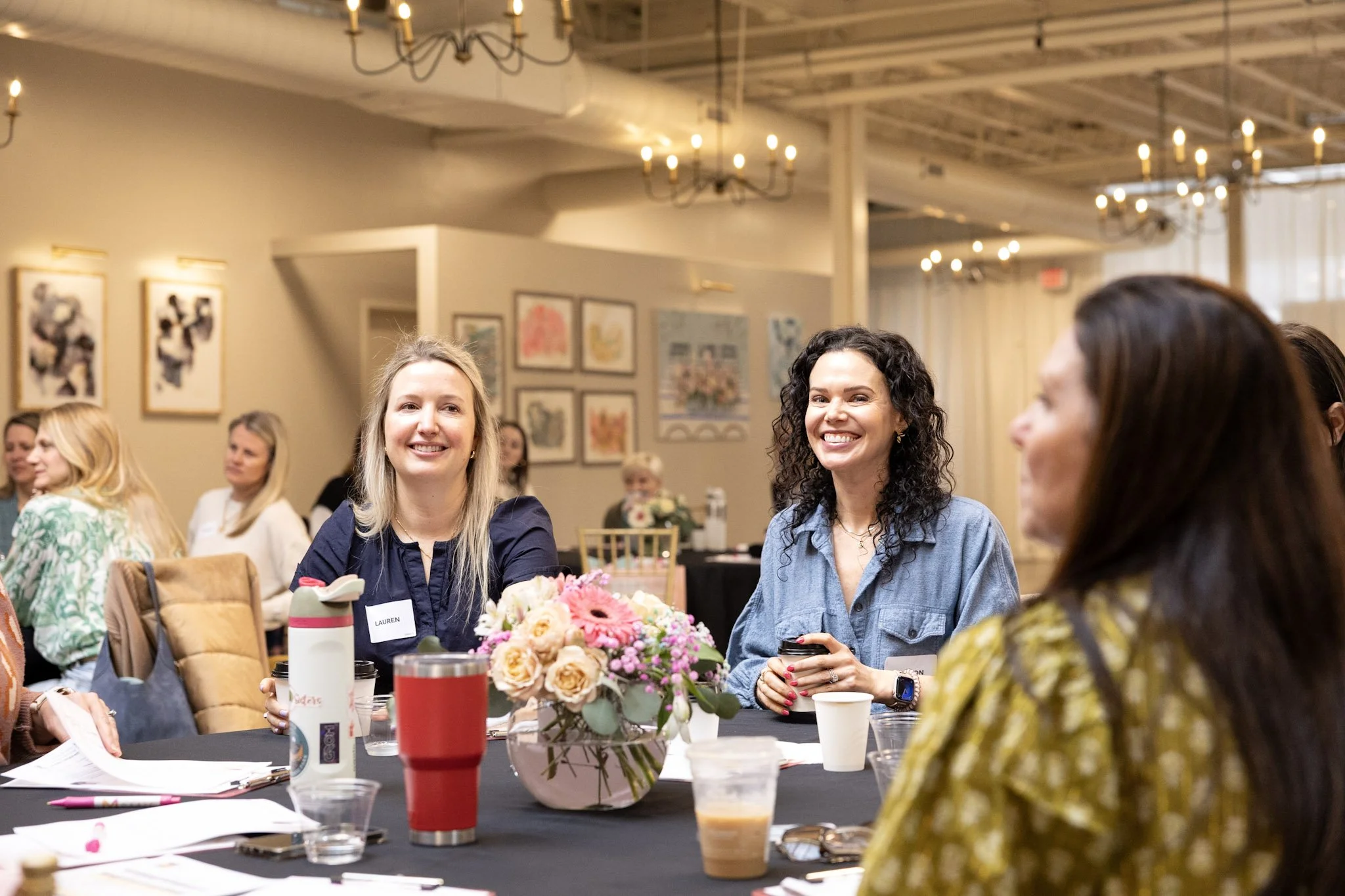 A group of women sitting around a table at a meeting or social event, with a floral centerpiece and drinks in front of them. The women are smiling and appear engaged. The room has artwork on the walls and chandeliers hanging from the ceiling.