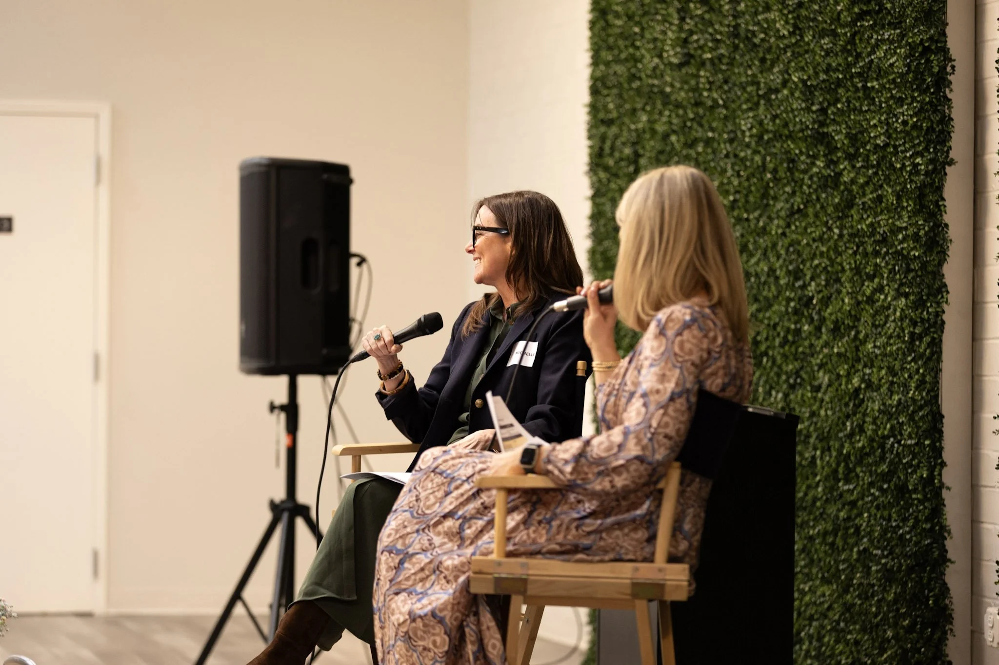 Two women sitting on chairs, participating in a panel discussion. The woman on the left is holding a microphone and smiling. The woman on the right is also holding a microphone. There is a large speaker to the left and a green wall with foliage behin