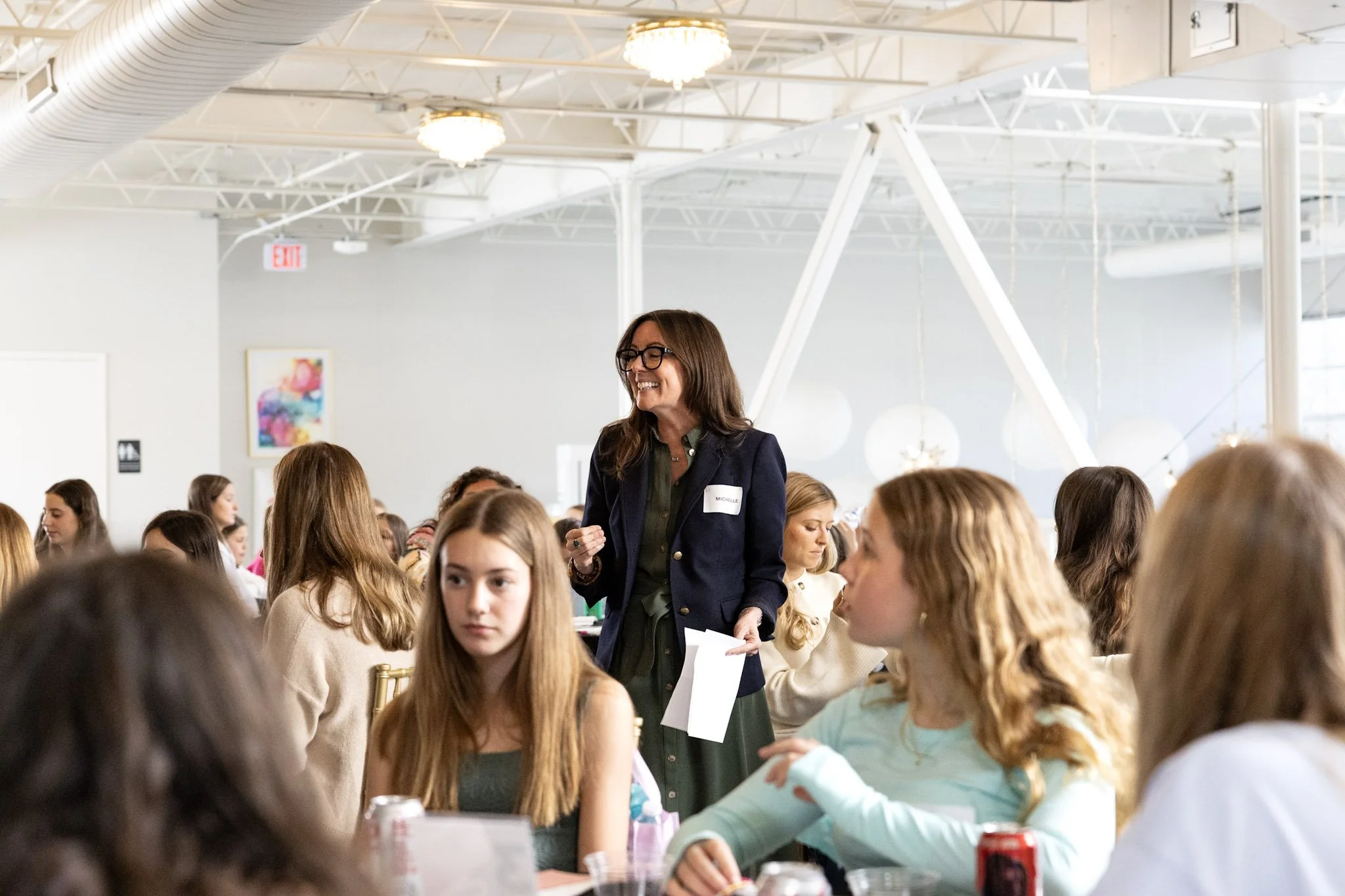A woman with glasses and long hair, wearing a dark blazer, standing and smiling, surrounded by young girls at a table in a bright, spacious room with high ceilings and white decor.