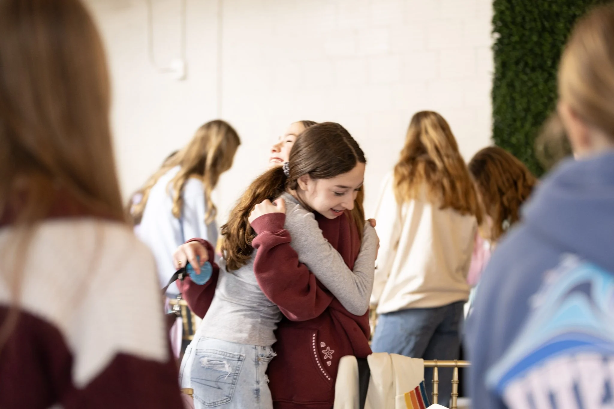 Young girl hugging another girl with eyes closed, surrounded by other people, in an indoor setting.