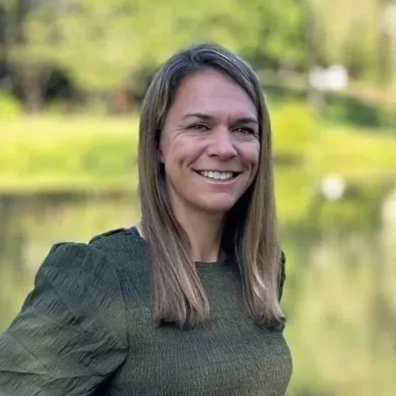 A woman with long brown hair smiling outdoors with a lake and green trees in the background.