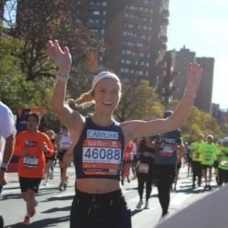 A woman running in a marathon, smiling and waving with both hands, surrounded by other runners in an outdoor city park or street with trees and buildings in the background.
