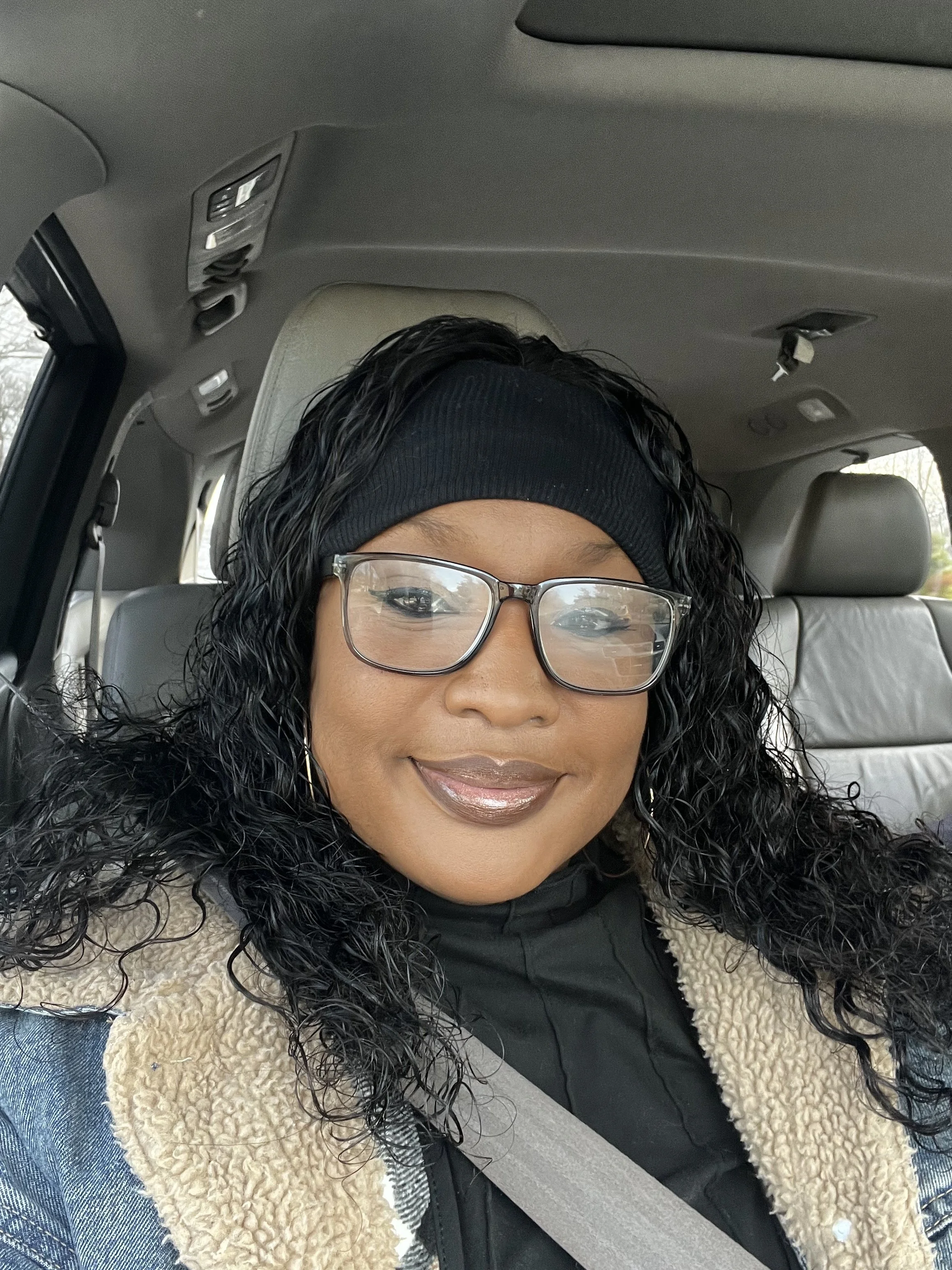A woman with curly black hair, glasses, and a black neck gaiter, smiling while sitting in a vehicle, wearing a black headband and a beige fleece jacket over a black shirt, with a seatbelt on.