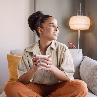 Young woman sitting on a couch, smiling, holding a mug, in a cozy living room with a lamp and flowers in the background.