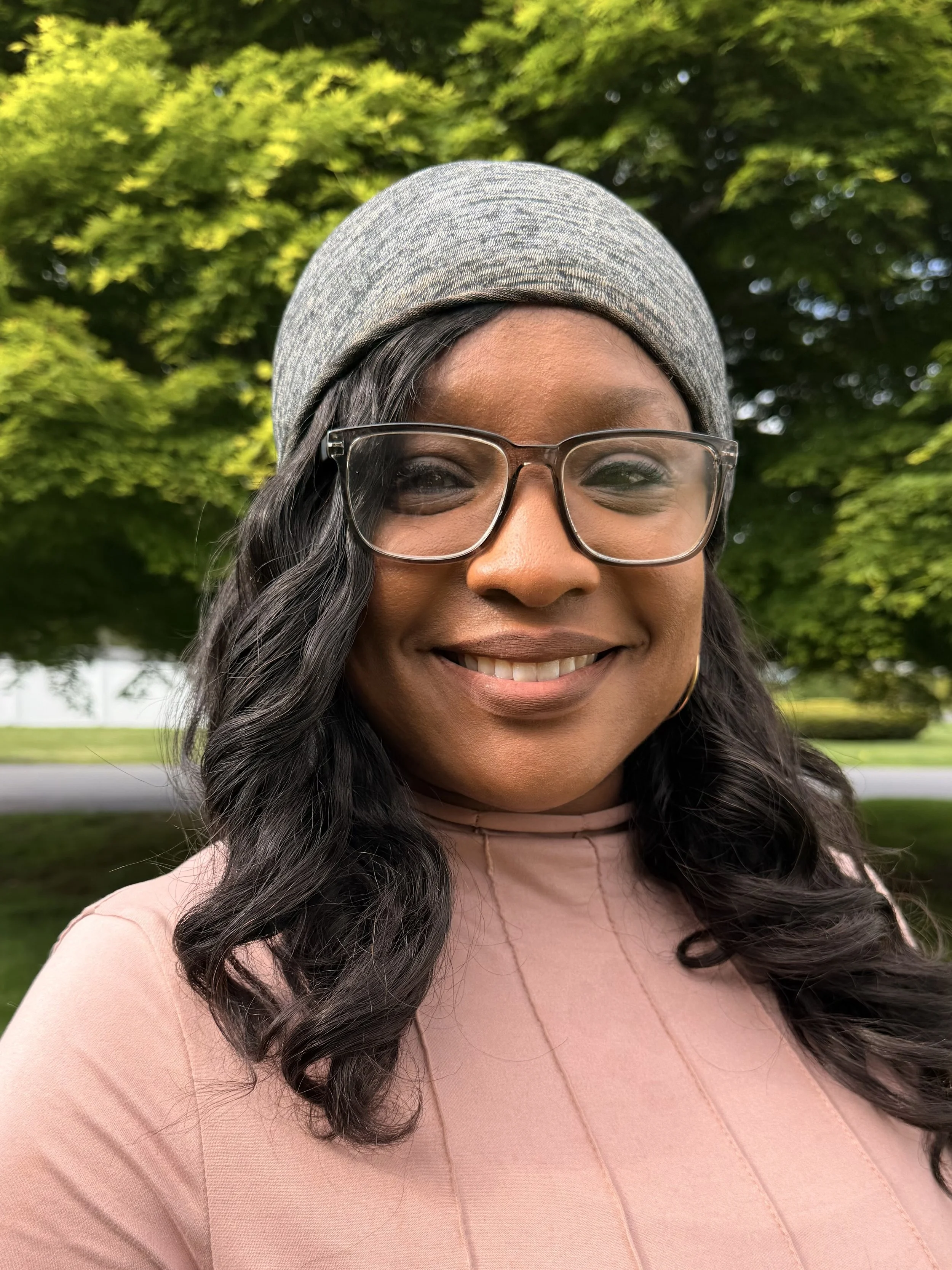 A smiling woman with glasses and a gray headscarf, outdoors with lush green trees in the background.