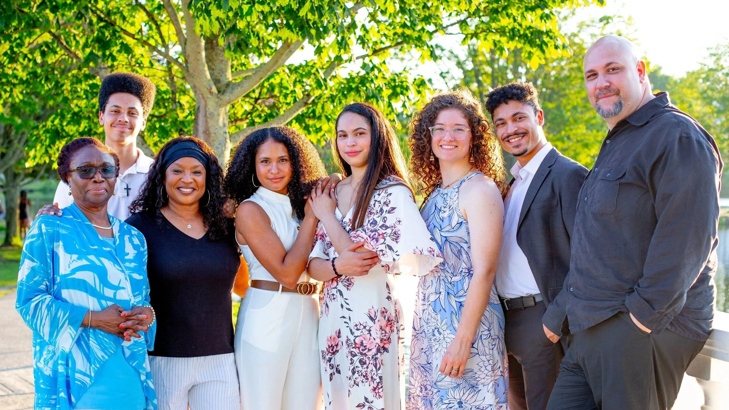 Group of ten diverse people standing outdoors in a park, smiling, with green trees in the background, during daytime.