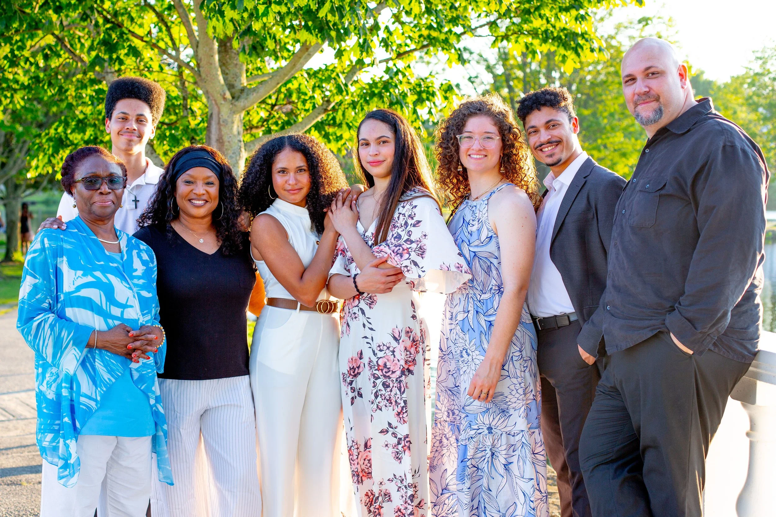 Group of diverse people smiling outdoors under a green leafy tree, dressed in casual and semi-formal clothing.
