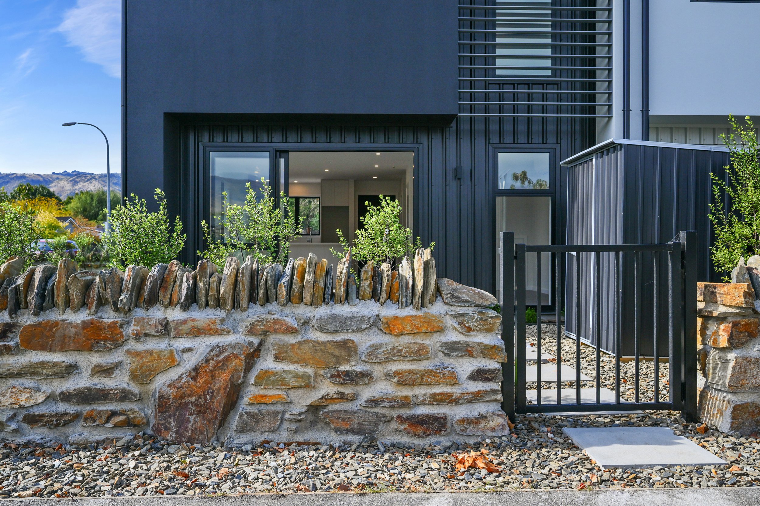 Schist stone boundary wall and entrance gate at Pisa Vistas, with contemporary dark-clad townhouse beyond.