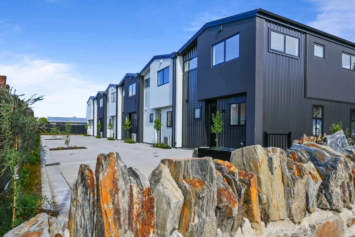 Row of Pisa Vistas townhouses viewed from street level, with schist boulders in the foreground and off-street parking alongside.