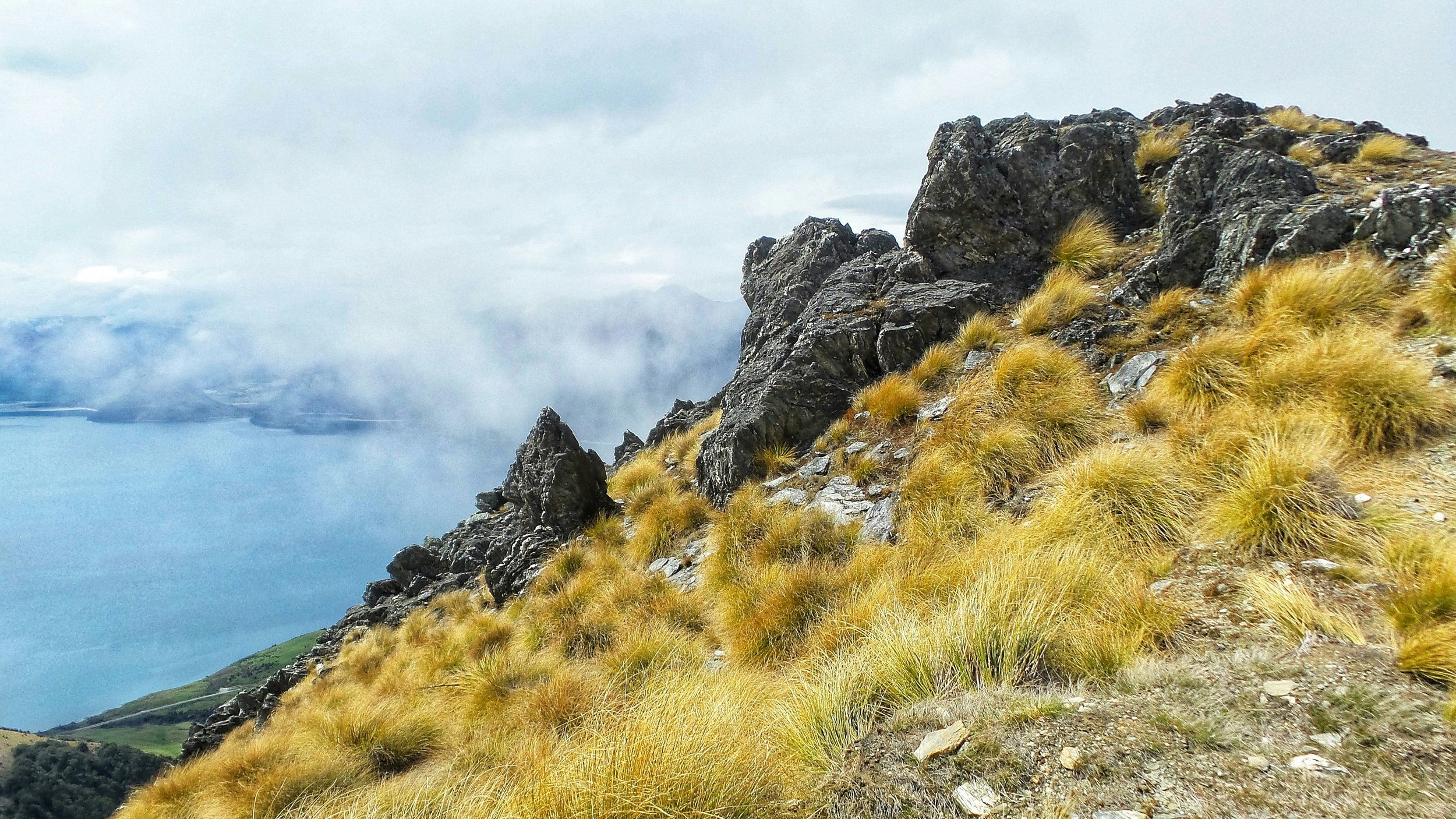 Rocky alpine ridge in Central Otago with tussock grass, jagged rock outcrops and cloud below.
