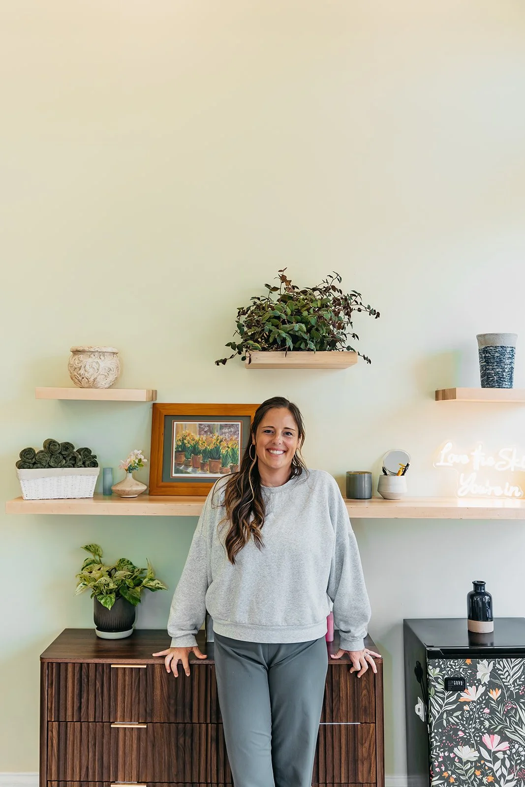 A woman with long brown hair, wearing a gray sweatshirt and gray pants, standing in front of a wooden sideboard and smiling at the camera inside a room with decorative shelves and plants on the wall.