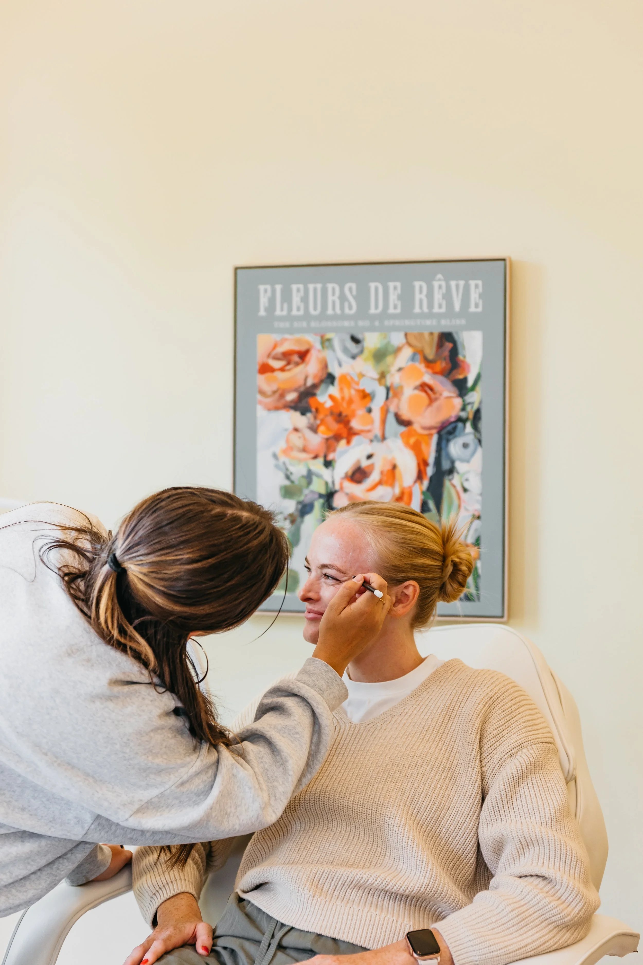 Makeup artist applying makeup to a woman seated in a chair, with a floral poster on the wall behind them.