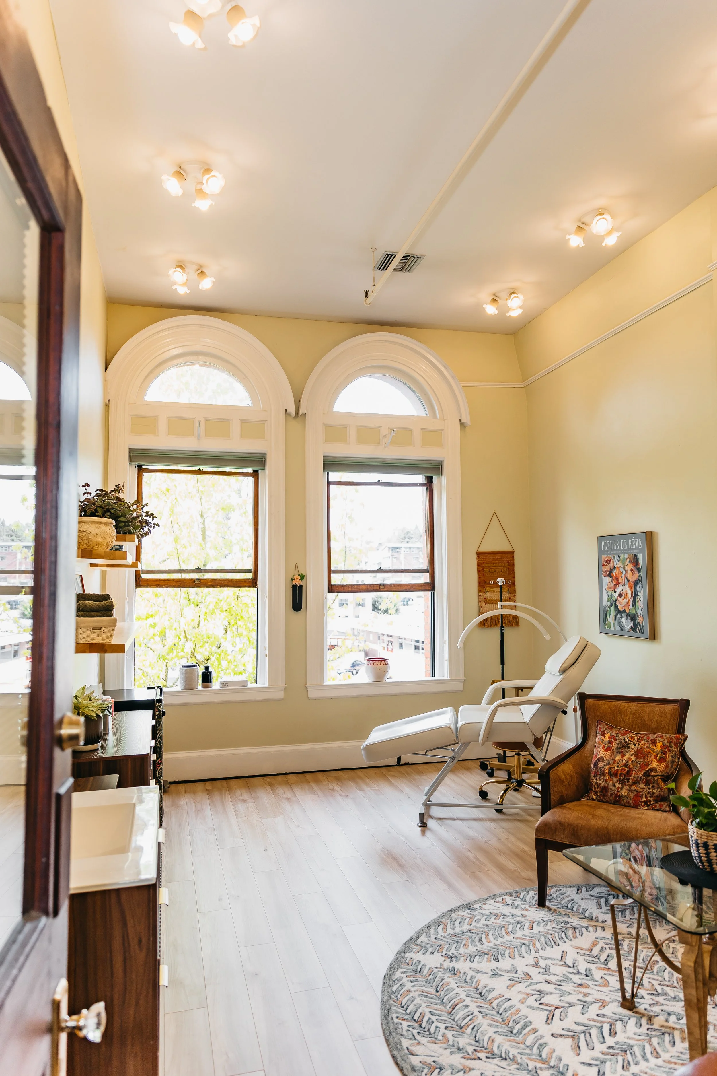 A room with light yellow walls and wooden flooring, featuring three large arched windows letting in natural light. There is a white reclining chair and a wooden armchair with a decorative pillow, a glass coffee table, and various plants and decor items on shelves and hung on the walls.
