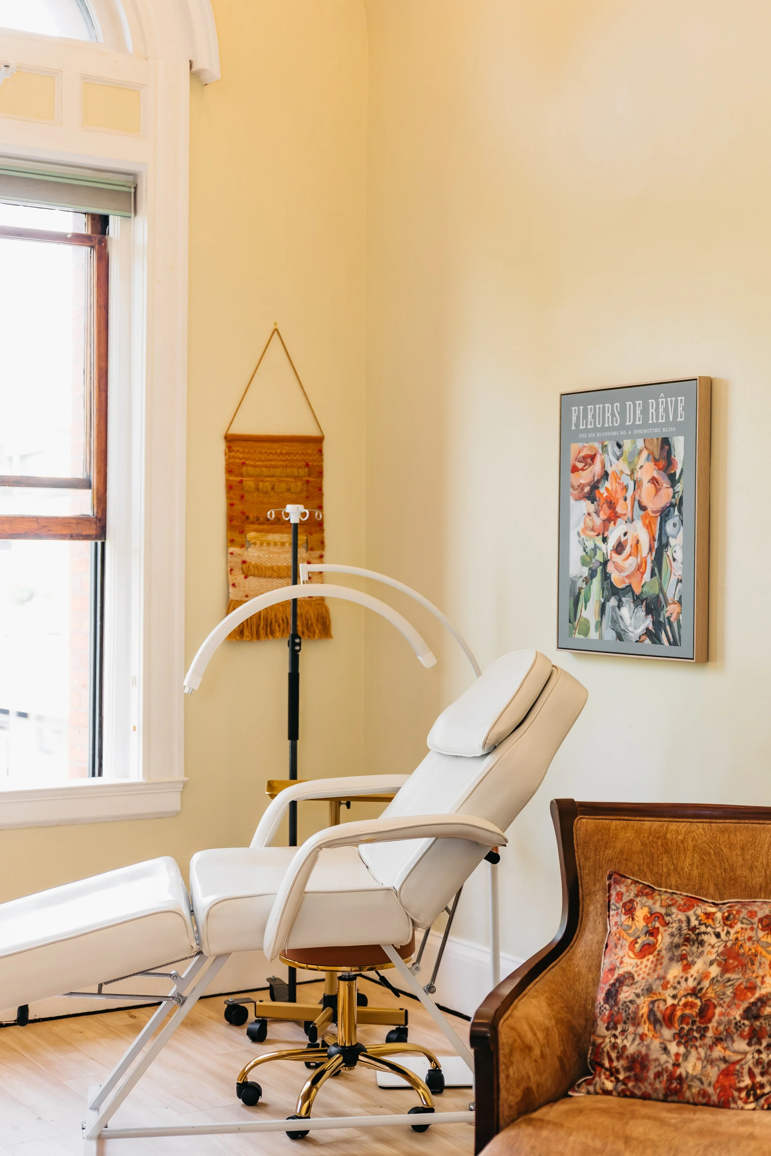 Medical examination room with a white adjustable chair, a medical lamp, a wooden chair with floral cushion, and framed botanical art on the beige wall near a window.