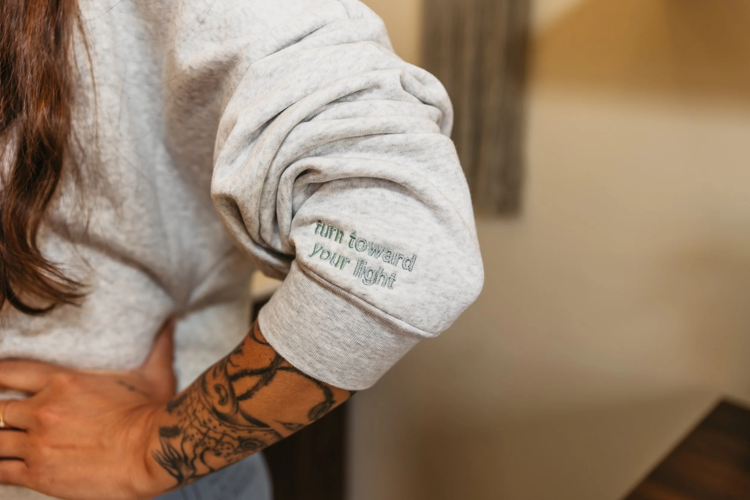 Close-up of a person wearing a light gray sweatshirt with rolled-up sleeves, revealing tattoos on their arm, holding a child. The child’s face is not visible.
