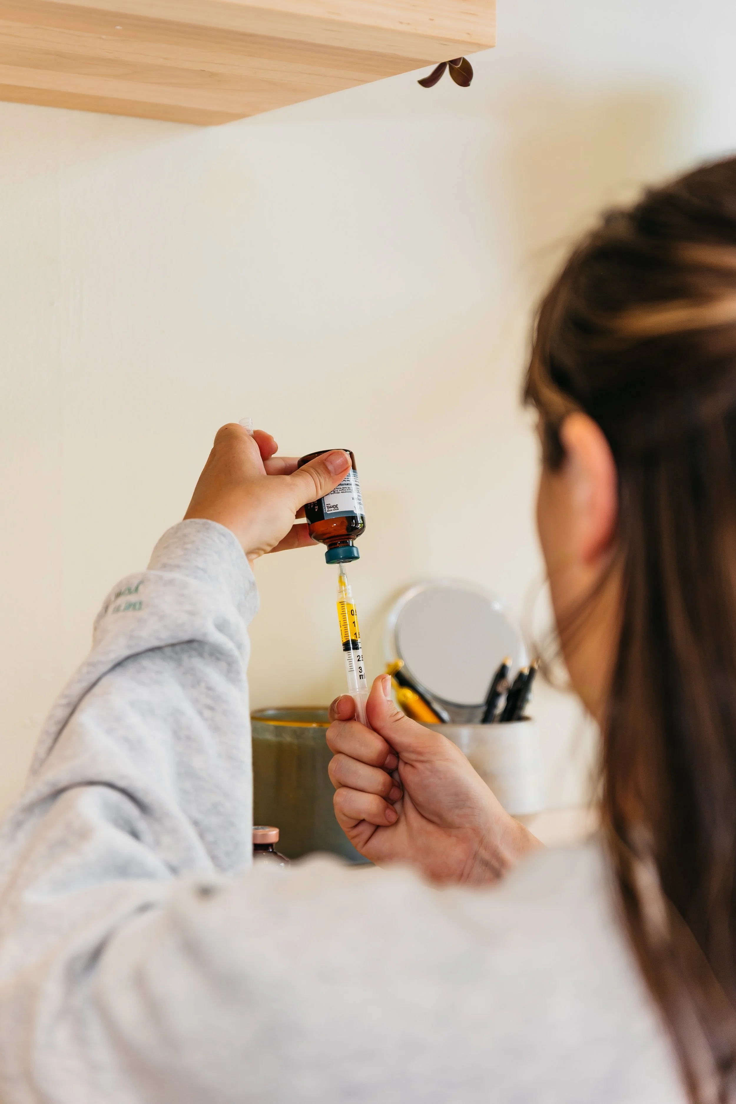 A person holding a small medical vial and a syringe, preparing for an injection, with another person's head partially visible, indoor setting with a shelf of pens or tools in the background.