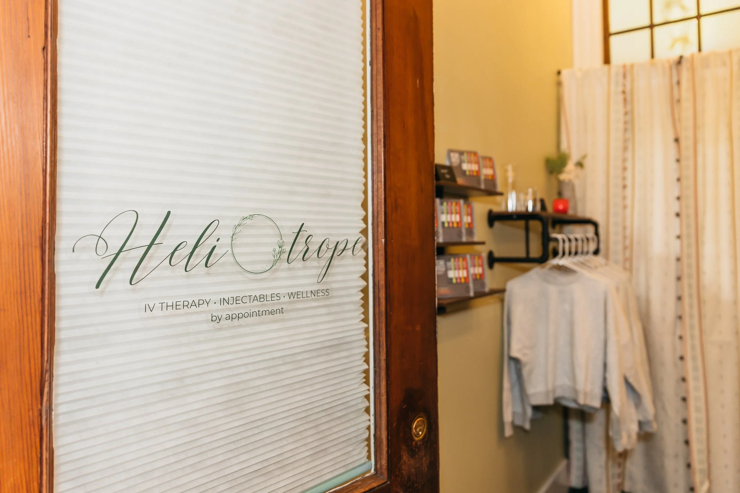 Close-up of a sign on a door that reads "Heli TROP" with additional text below mentioning IV therapy, injectables, and wellness by appointment. The background shows a reception area with shelves, books, and a chair.