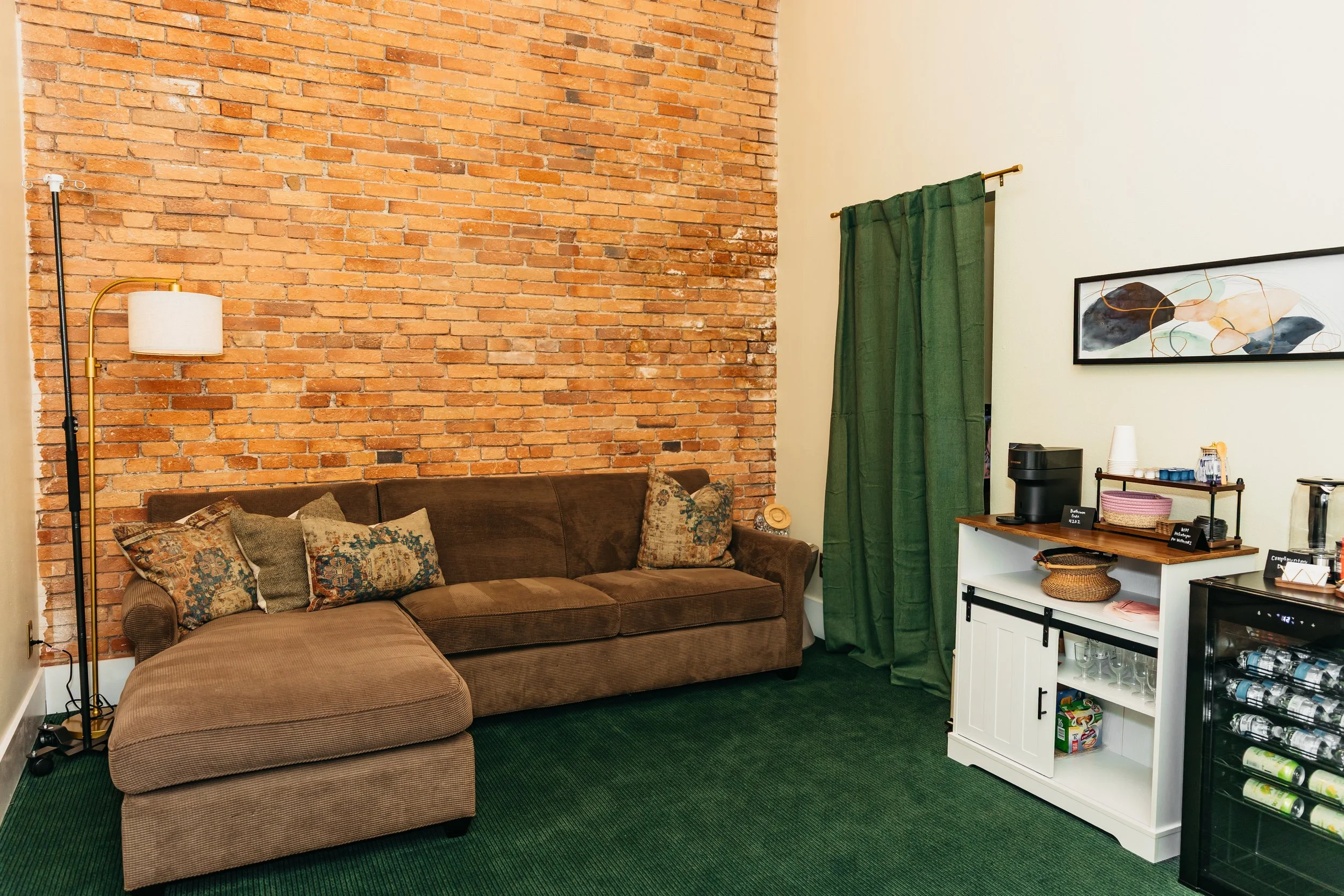 Living room with a brown upholstered sectional sofa and decorative pillows, a red brick accent wall, a floor lamp with a white shade and brass arm, a white cabinet with a coffee maker and glassware, a framed abstract art piece on the wall, and green curtains covering a window.