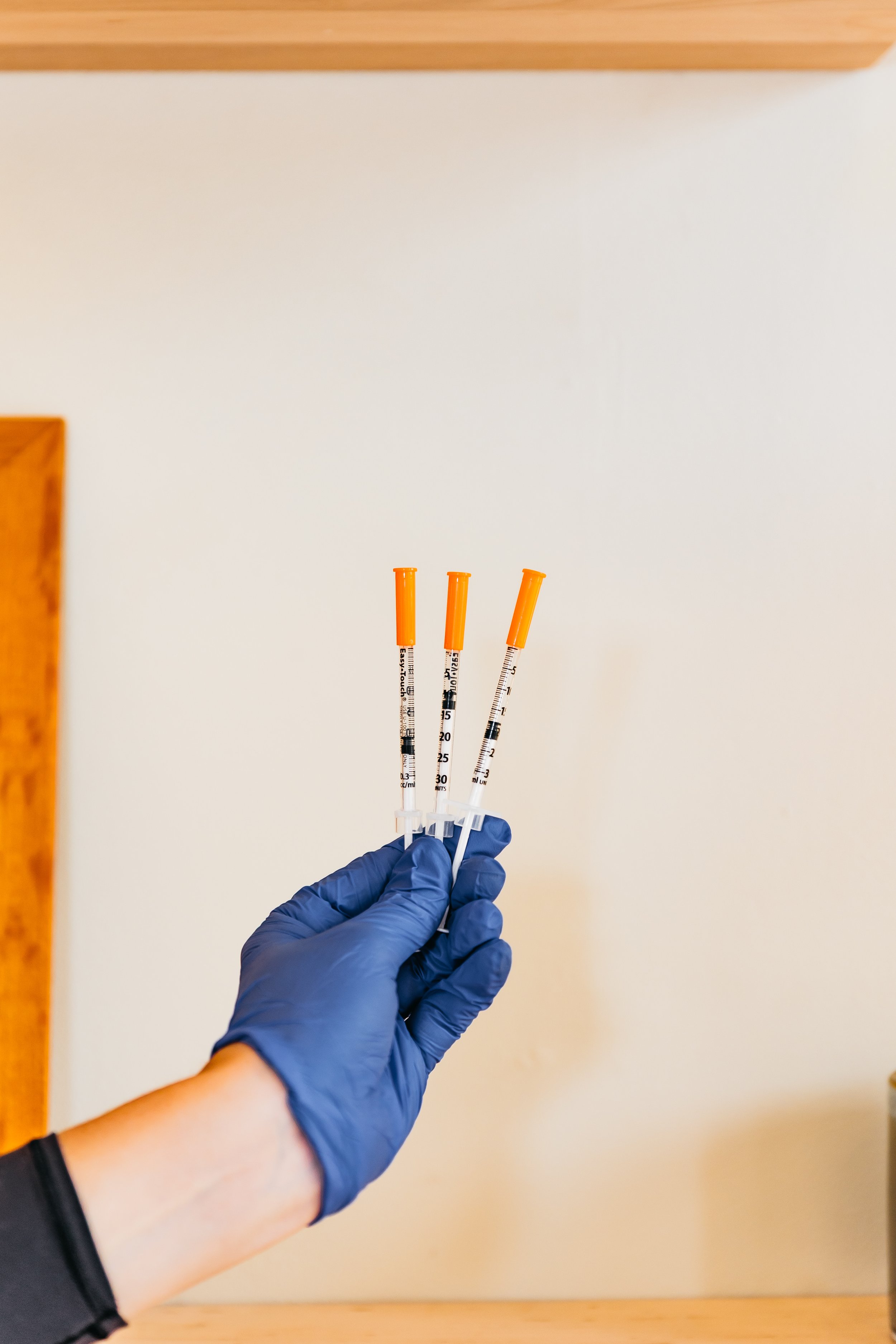 A person wearing a blue glove holding three syringes with orange caps and measurement markings against a background of a white wall and wooden corner.