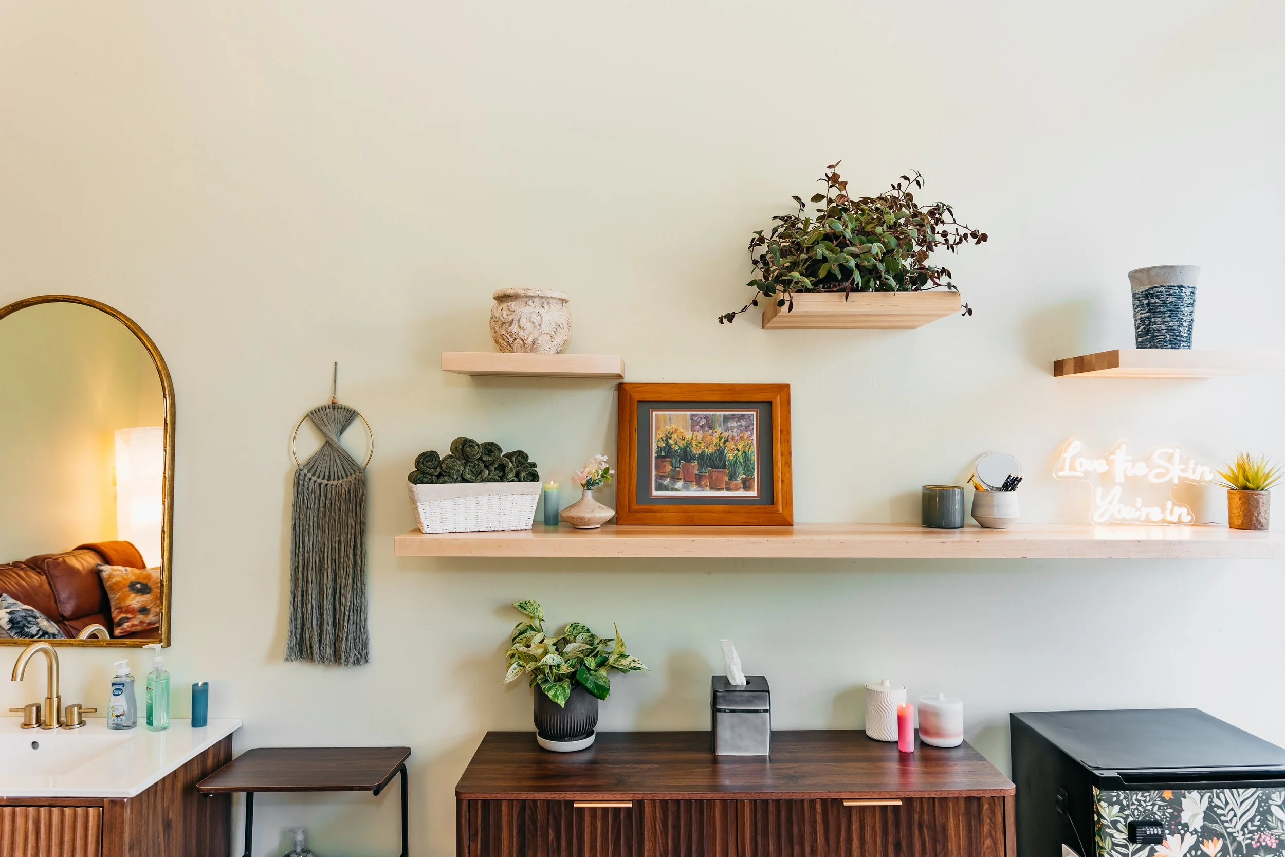 Decorative wall with floating shelves holding various potted plants, framed pictures, and ornaments. A salon or spa workspace with bottles, candles, and a sign that reads 'Love to Skin You're In'.