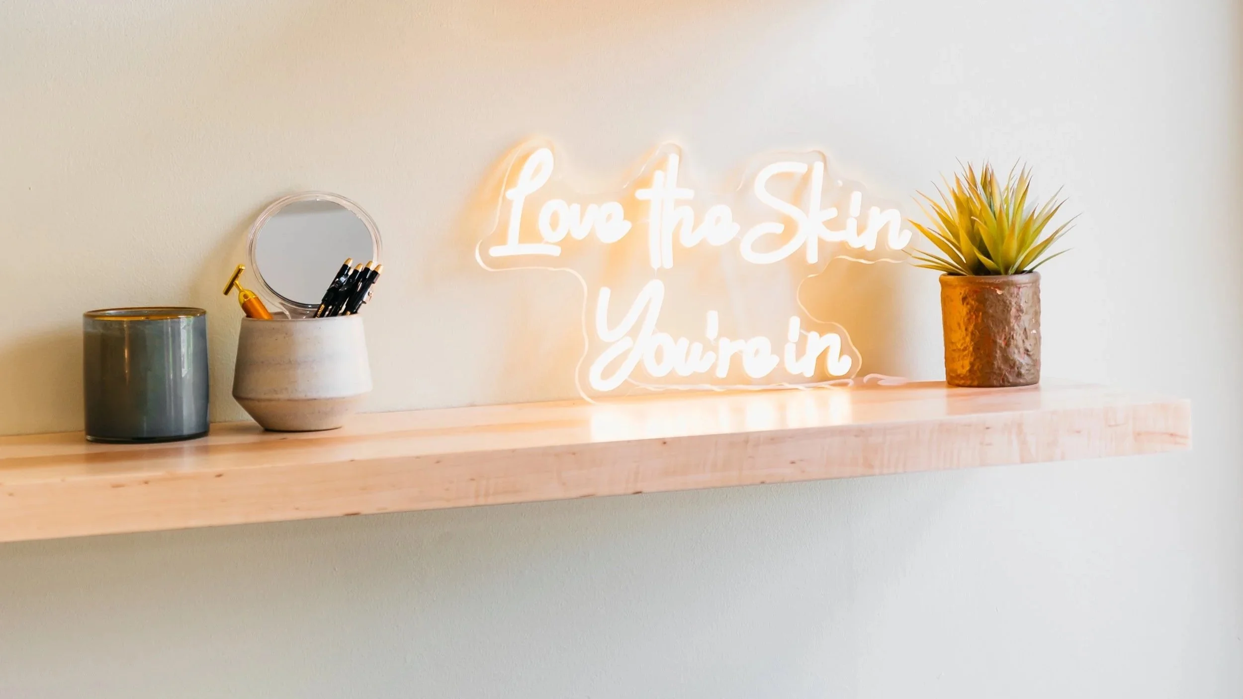 Decorative shelf with a small round mirror, a candle in a jar, a white pencil holder with black pens, a yellow-green potted plant, and a neon sign reading 'Love the Skin You're In'