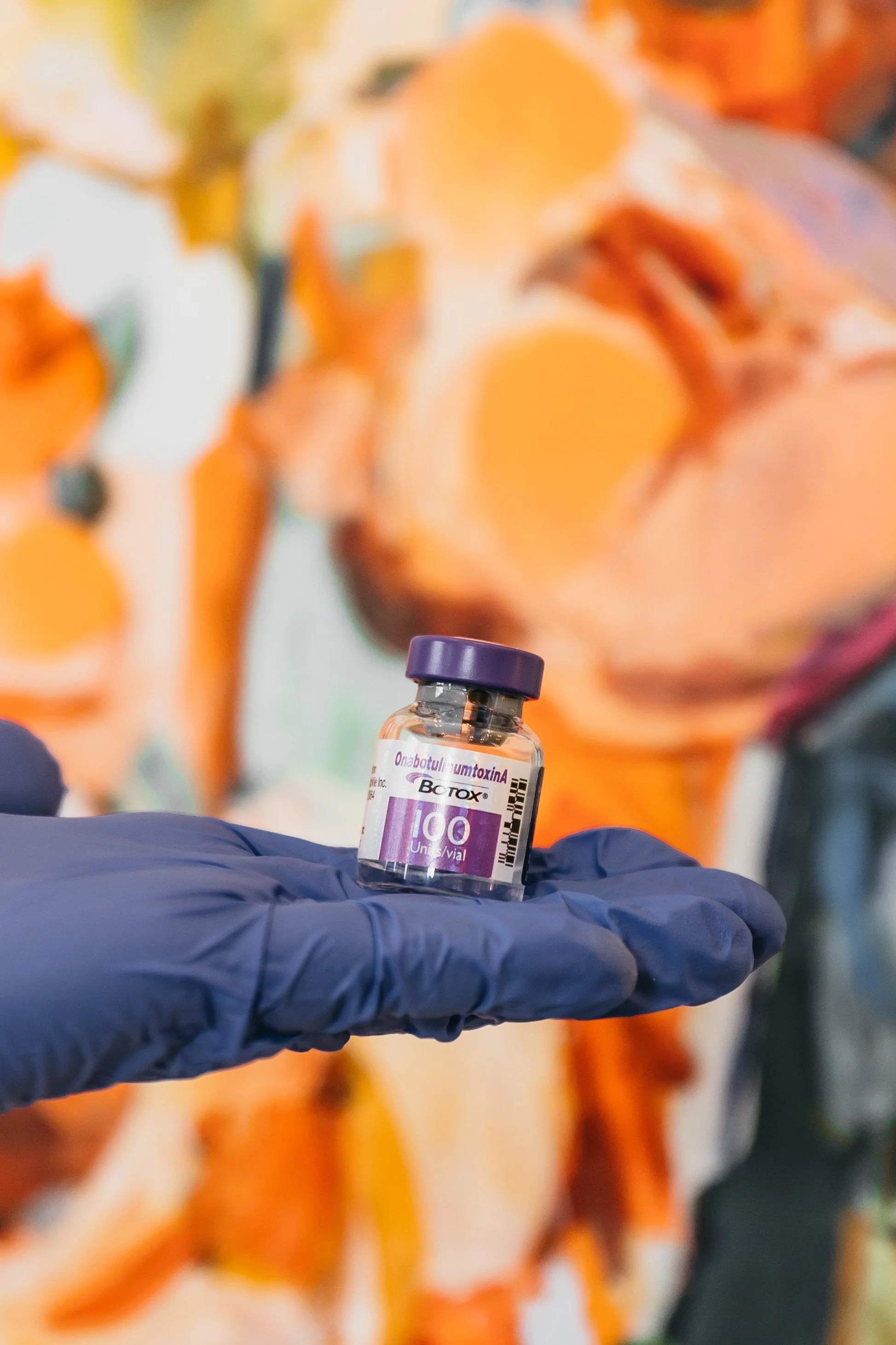 A gloved hand holding a small vial labeled 'Botox' with a colorful background featuring blurry orange artwork.