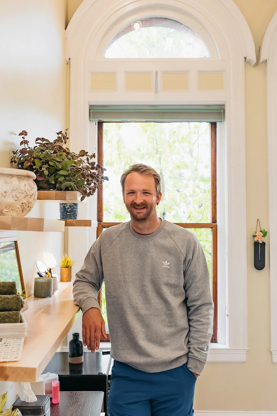 A man with a beard and brown hair smiling while standing in a bright room with large window behind him. He is wearing a gray Adidas sweatshirt and blue pants, leaning on a wooden countertop with plants, a mirror, and decor items.