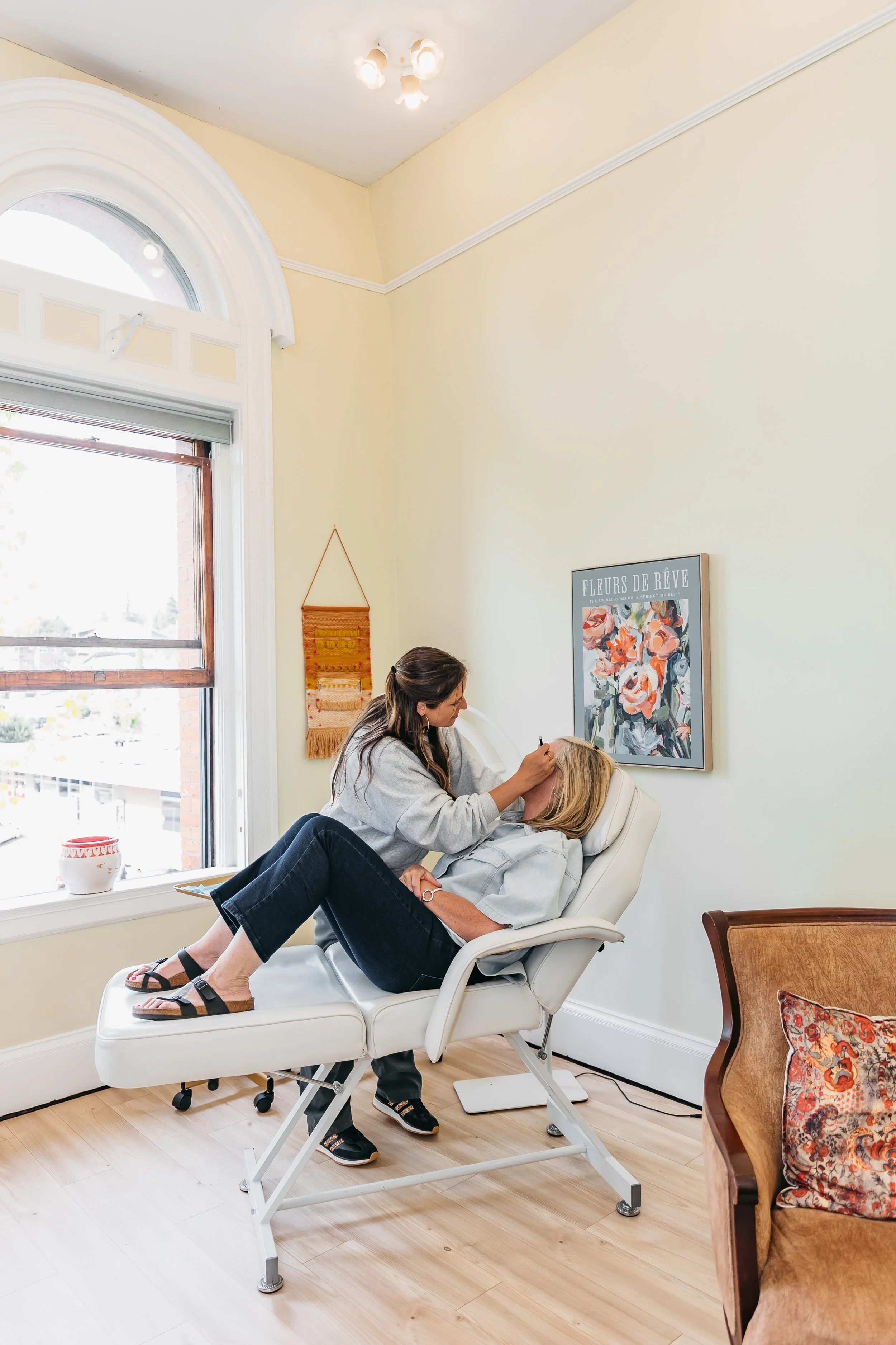 A woman with long brown hair dressed in a grey hoodie and black sandals is getting a facial or beauty treatment from a woman with blonde hair, reclining in a white treatment chair with her eyes covered with a white pillow. The scene takes place in a bright room with large window, framed art, and warm decor.