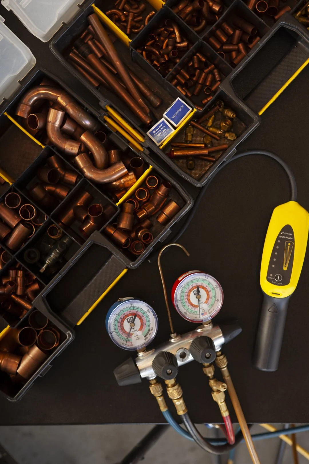 A collection of copper plumbing fittings in plastic storage bins, along with a refrigerant manifold gauge set and a yellow infrared thermometer on a black work surface.