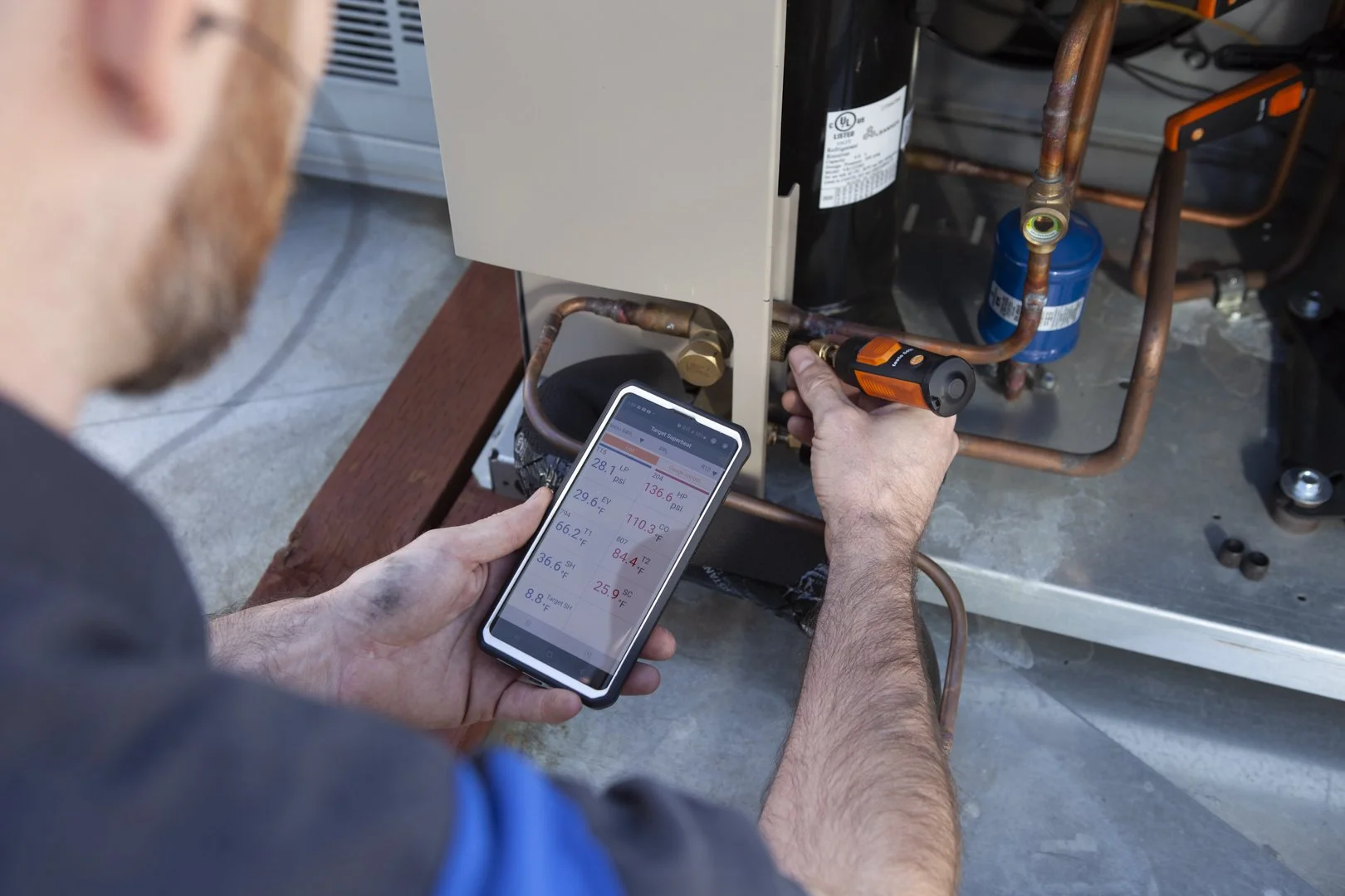 A technician checks air conditioning or refrigeration system pressures and temperatures using a digital manifold gauge app on a smartphone while holding a temperature sensor or probe, with copper pipes and a compressor in the background.