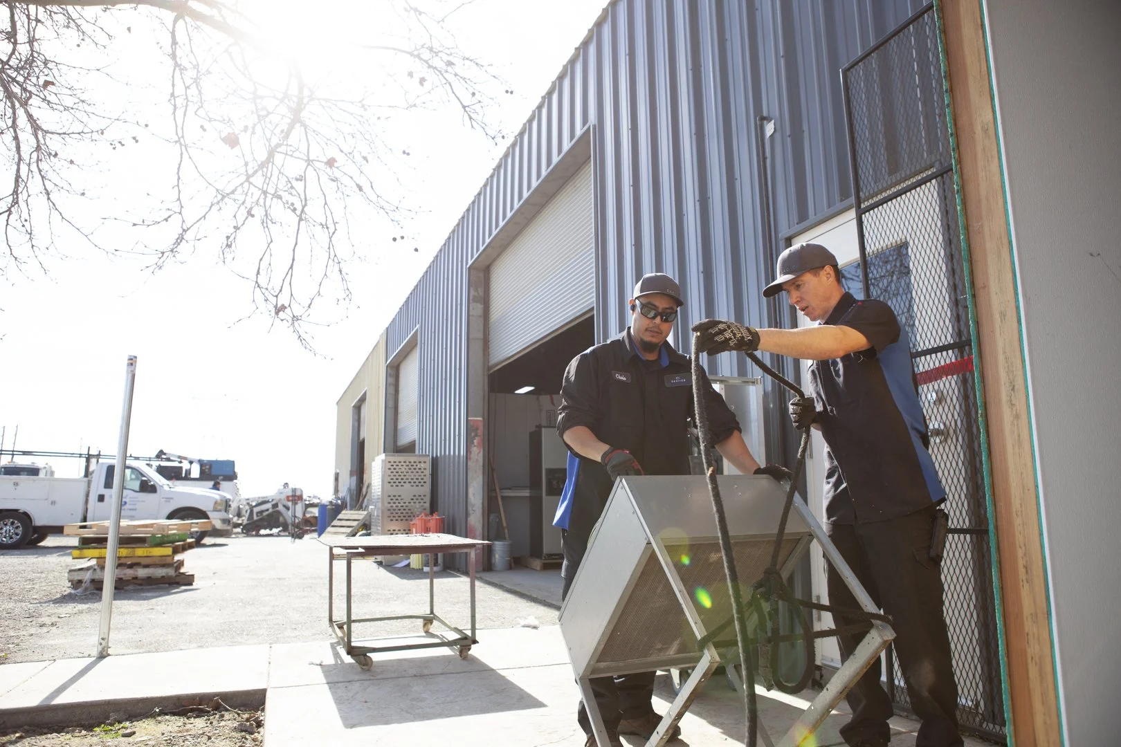 Two construction workers wearing black uniforms and caps working outside a metal warehouse. One worker is holding a wire while the other is handling a piece of equipment. Construction tools and vehicles are visible in the background, and the sun is shining brightly.