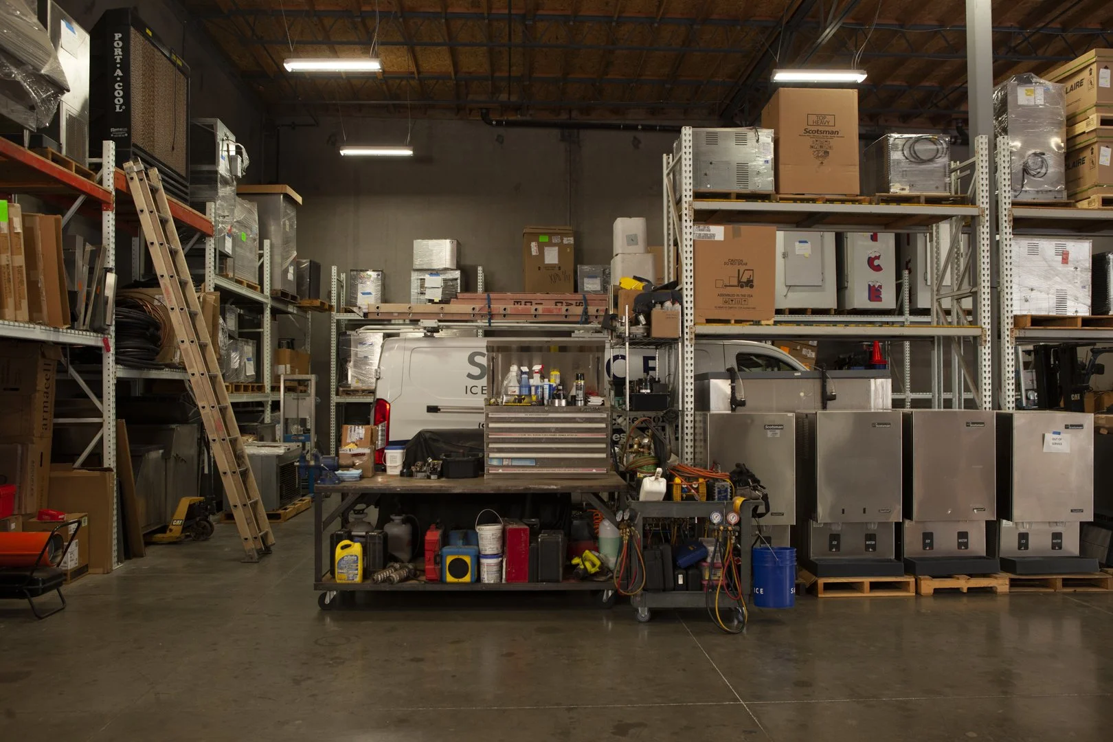 Industrial warehouse with shelves filled with boxes, tools, and equipment for refrigeration and appliance repair, including a white van and stainless steel machinery.
