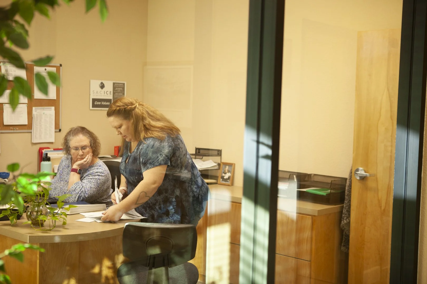 Two women are working at a desk in an office, with papers and a plant on the desk, and a bulletin board on the wall behind them.