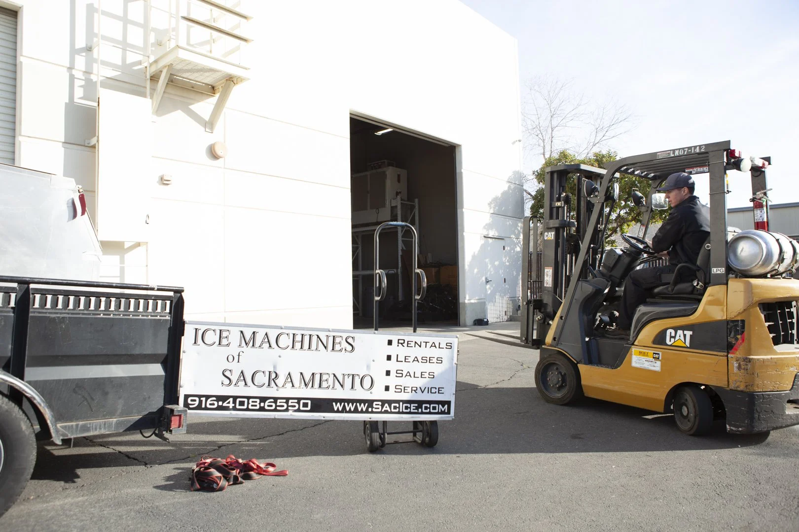 A forklift operator drives a yellow CAT forklift with a propane tank attached, near a white building. A sign in front of the building reads 'Ice Machines of Sacramento' and lists services such as rentals, leases, sales, and service, along with a phone number and website. There are some straps on the ground and a trailer partially visible on the left.