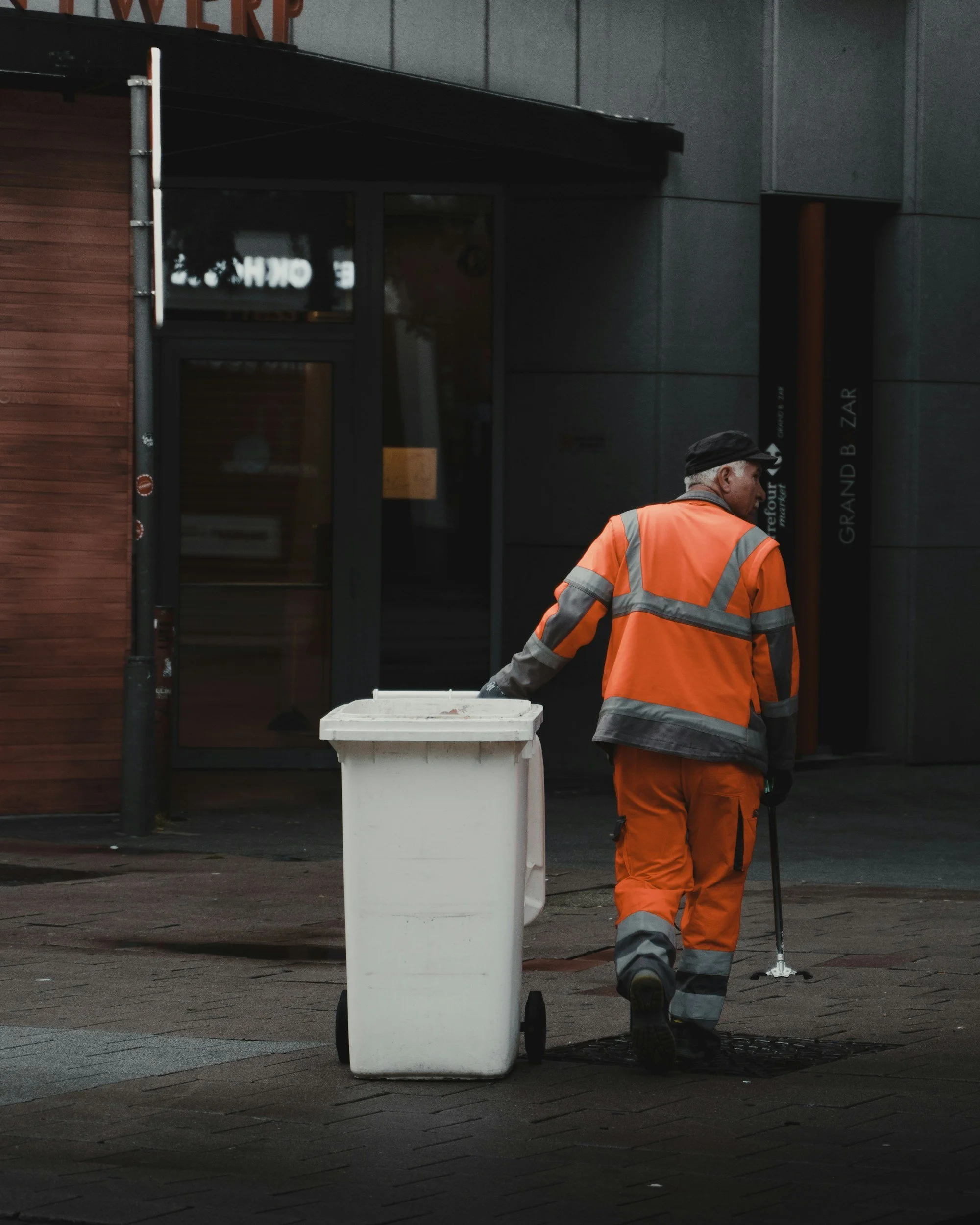 A street worker in an orange reflective uniform walking with a white trash bin on wheels in front of a dark building with wooden and gray walls.