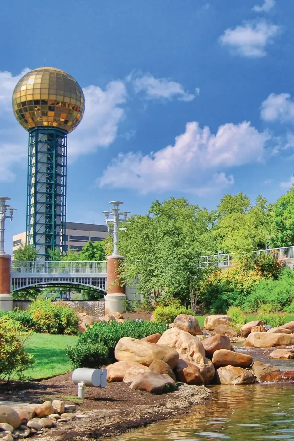 The image shows a park in Knoxville, TN with a rocky stream in the foreground, and a large spherical gold-colored structure on a tall blue tower behind lush green trees under a partly cloudy blue sky.