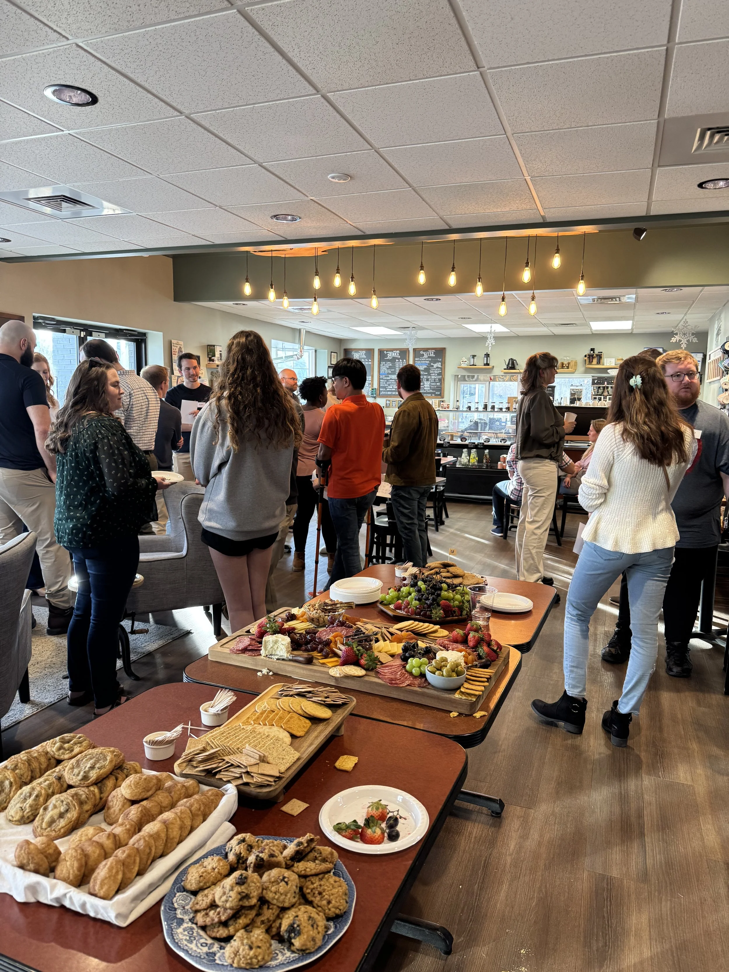 A gathering of people in a dining area with a buffet table of assorted baked goods, cookies, fruits, and snacks, under warm lighting fixtures.