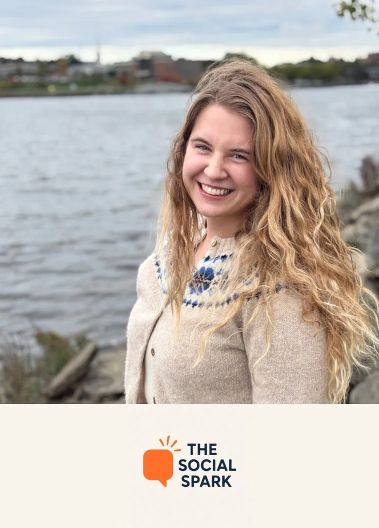 A smiling young woman with curly blonde hair stands outdoors near a river, wearing a beige knit sweater with a blue pattern, with city buildings in the background and the logo of The Social Spark at the bottom.